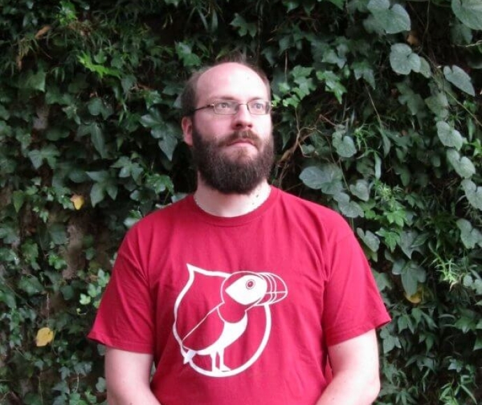Un homme barbu avec des lunettes et une forte calvitie porte un t-shirt rouge du drupalcamp lannion 2017, son expression est neutre, il regarde au loin