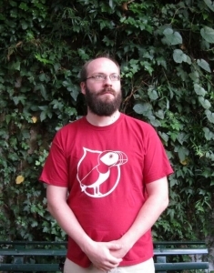 Un homme barbu avec des lunettes et une forte calvitie porte un t-shirt rouge du drupalcamp lannion 2017, son expression est neutre, il regarde au loin